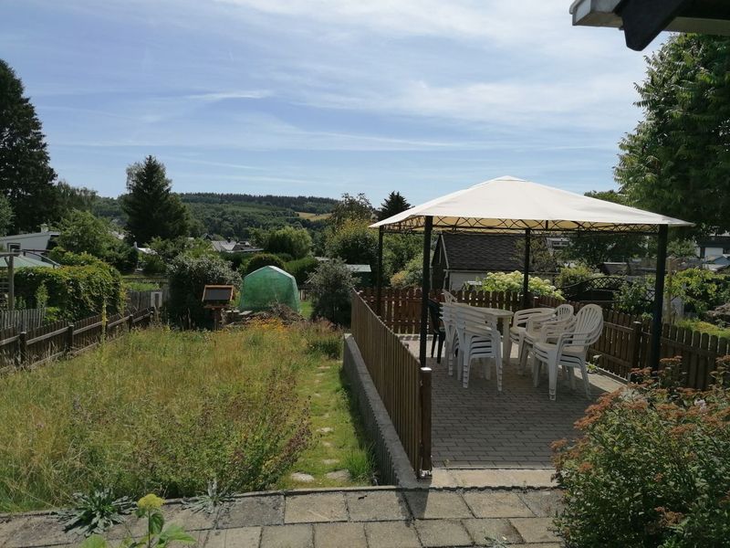 Mehrgenerationengarten mit Pavillon und weißer Sitzgruppe. Im Vordergrund sind blühende Sträucher zu sehen, die zusammen mit der Wildwiese ein naturnahes Ambiente schaffen.
