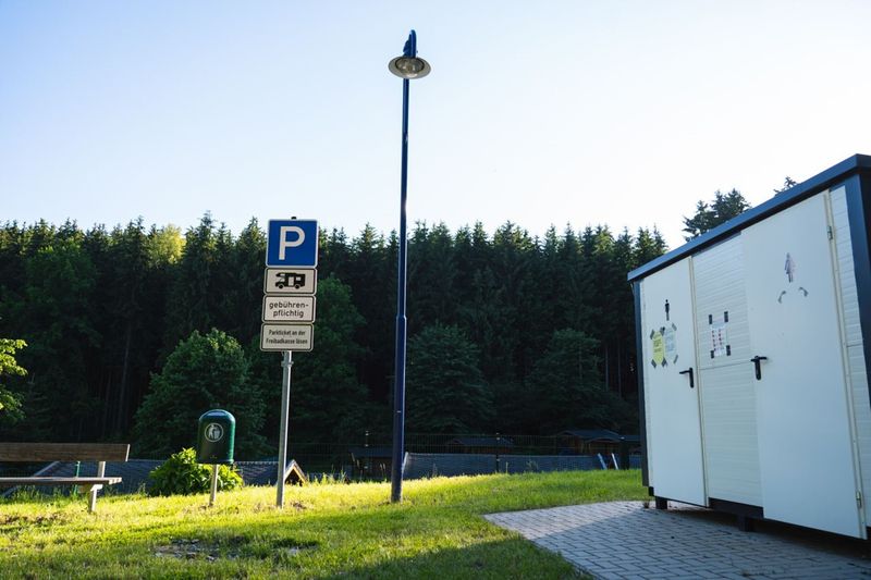 Query successful Ein markiertes Parkplatzschild für Wohnmobile vor einem dichten Waldpanorama und einer Sanitäranlage. (Link öffnet das Foto vergrößert in einem Popupfenster)