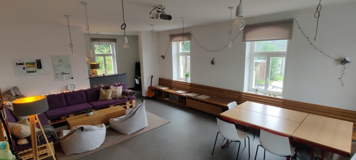 Heller Gemeinschaftsraum mit lila Palettensofa, Sitzsack, langen Holzbänken, Tischen und Beamer. (Bildlink öffnet sich vergrößert in Popup-Fenster)